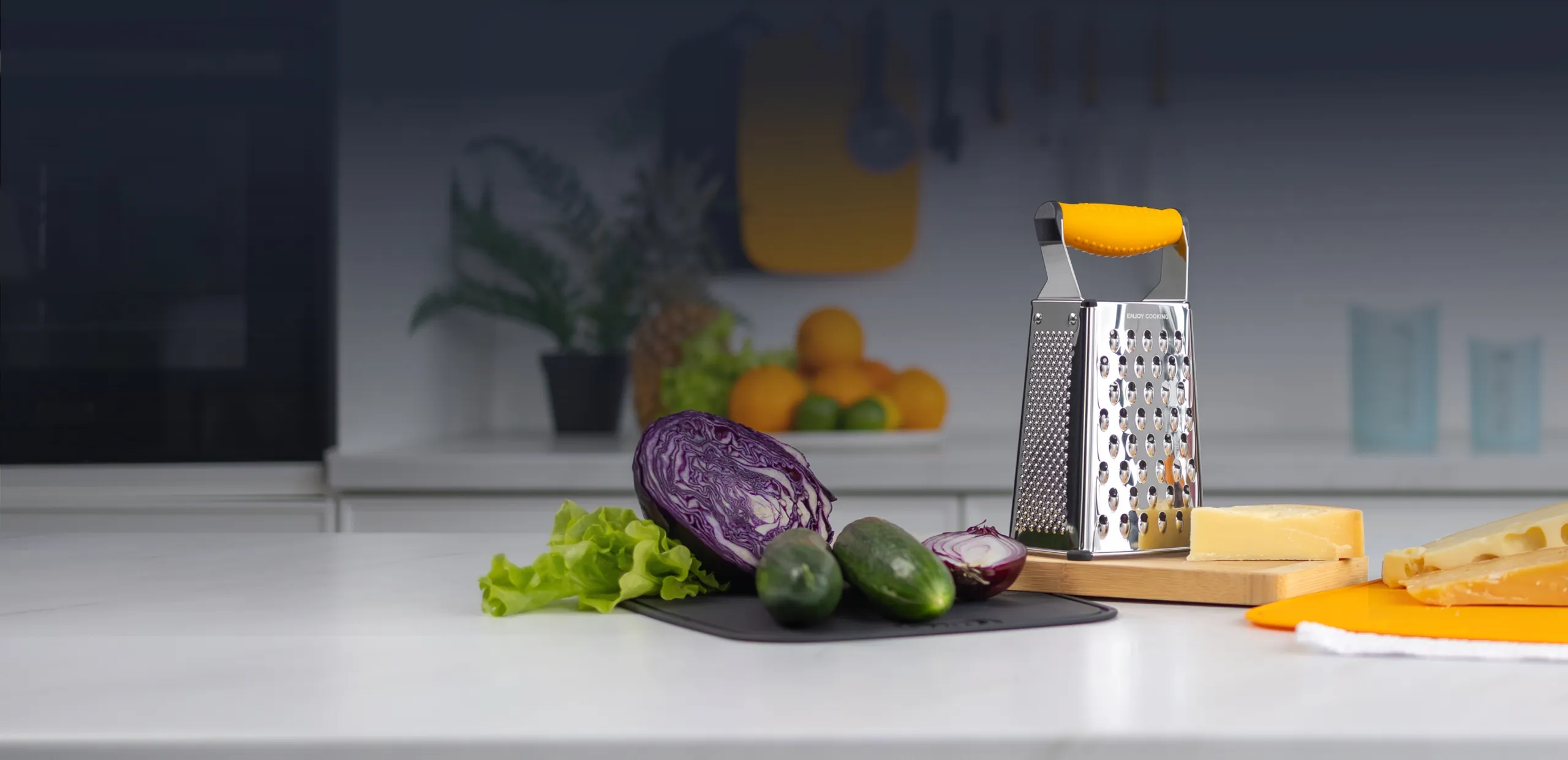 Deiss PRO box grater with a yellow handle on a kitchen counter, next to cheese and fresh vegetables